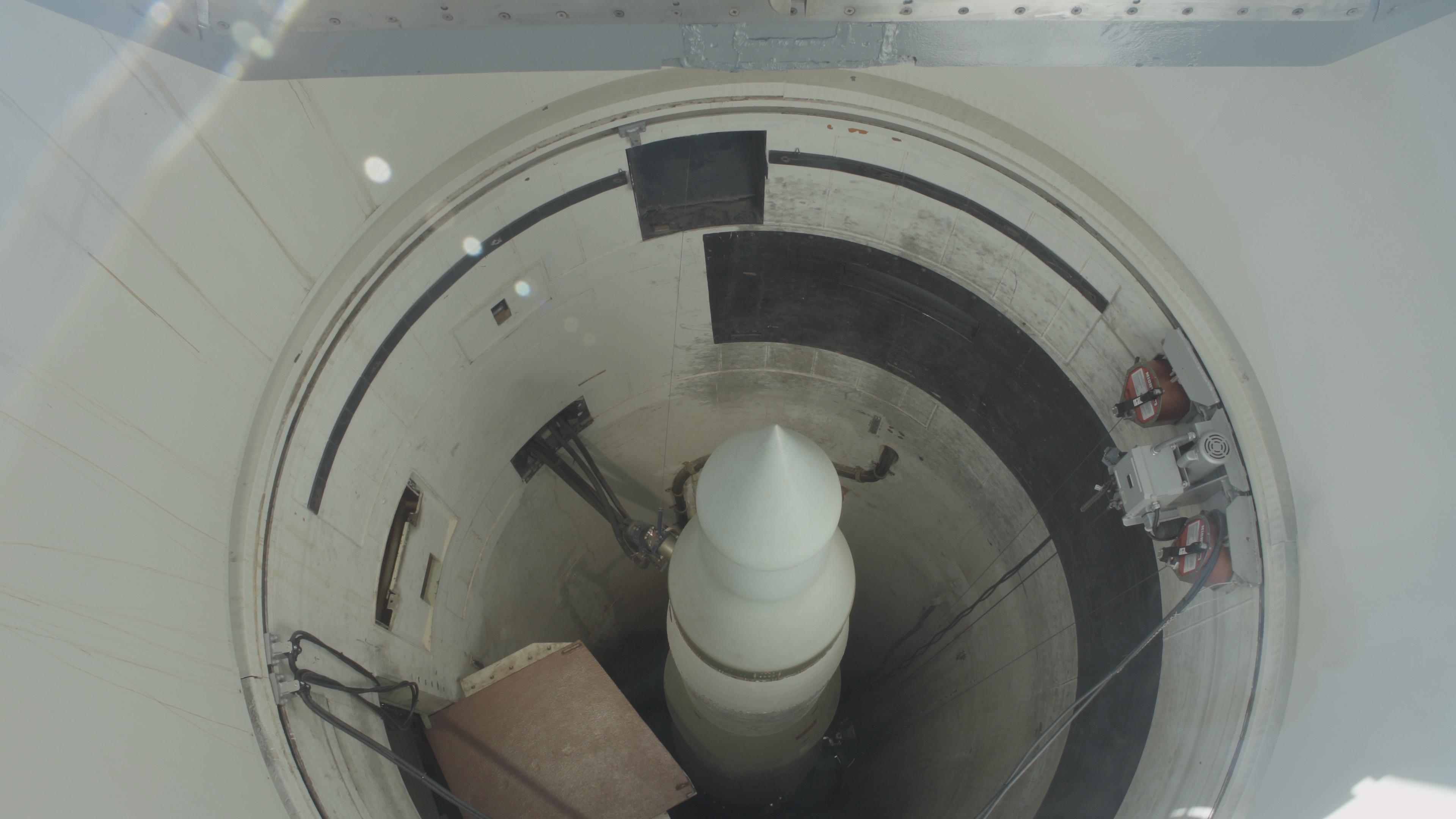 An overhead view looks down into a cylindrical missile silo containing a white training missile.