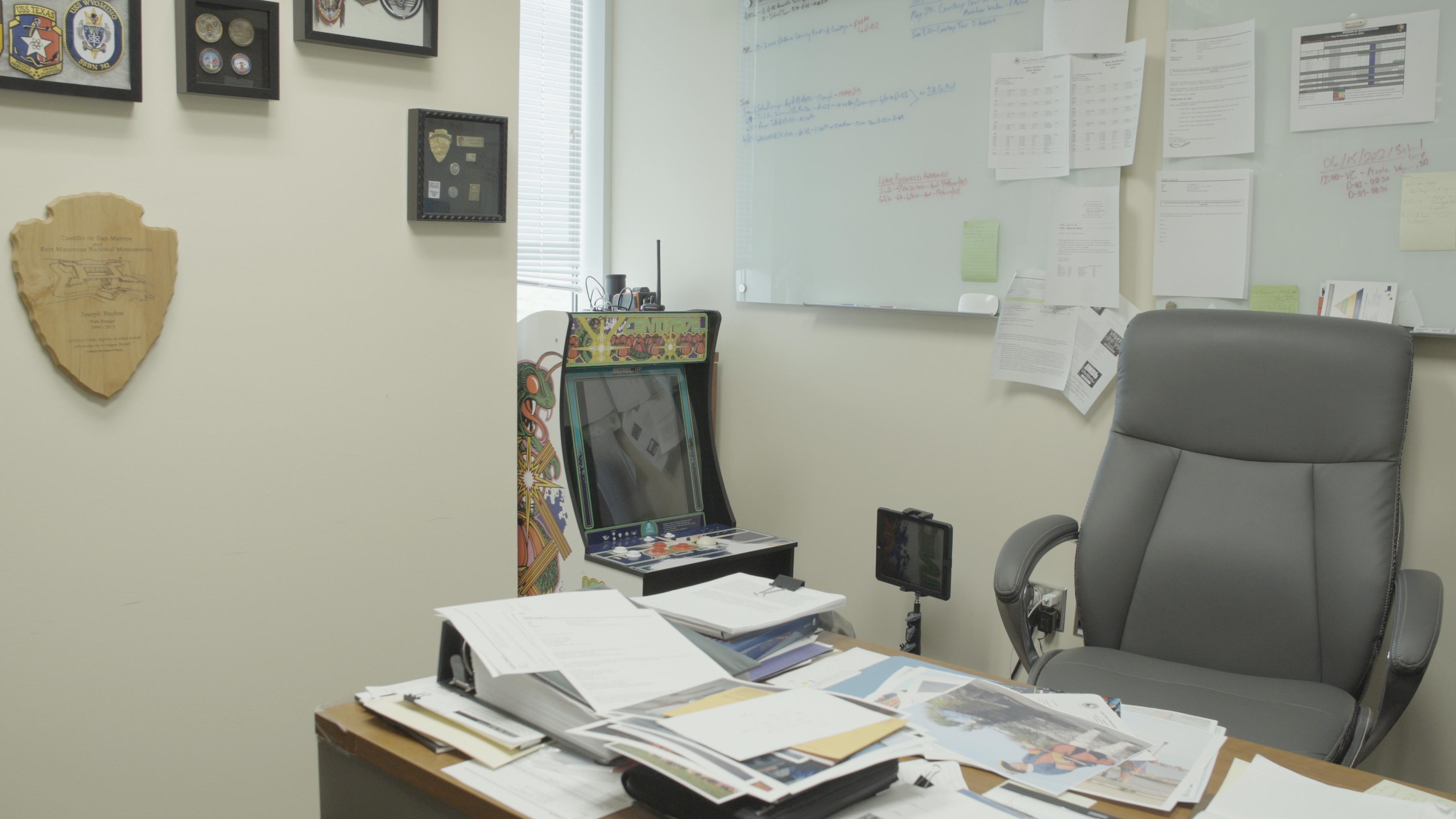 An Atari console and joystick sit on a desk in the park office, surrounded by maps, notes, and National Park Service material.