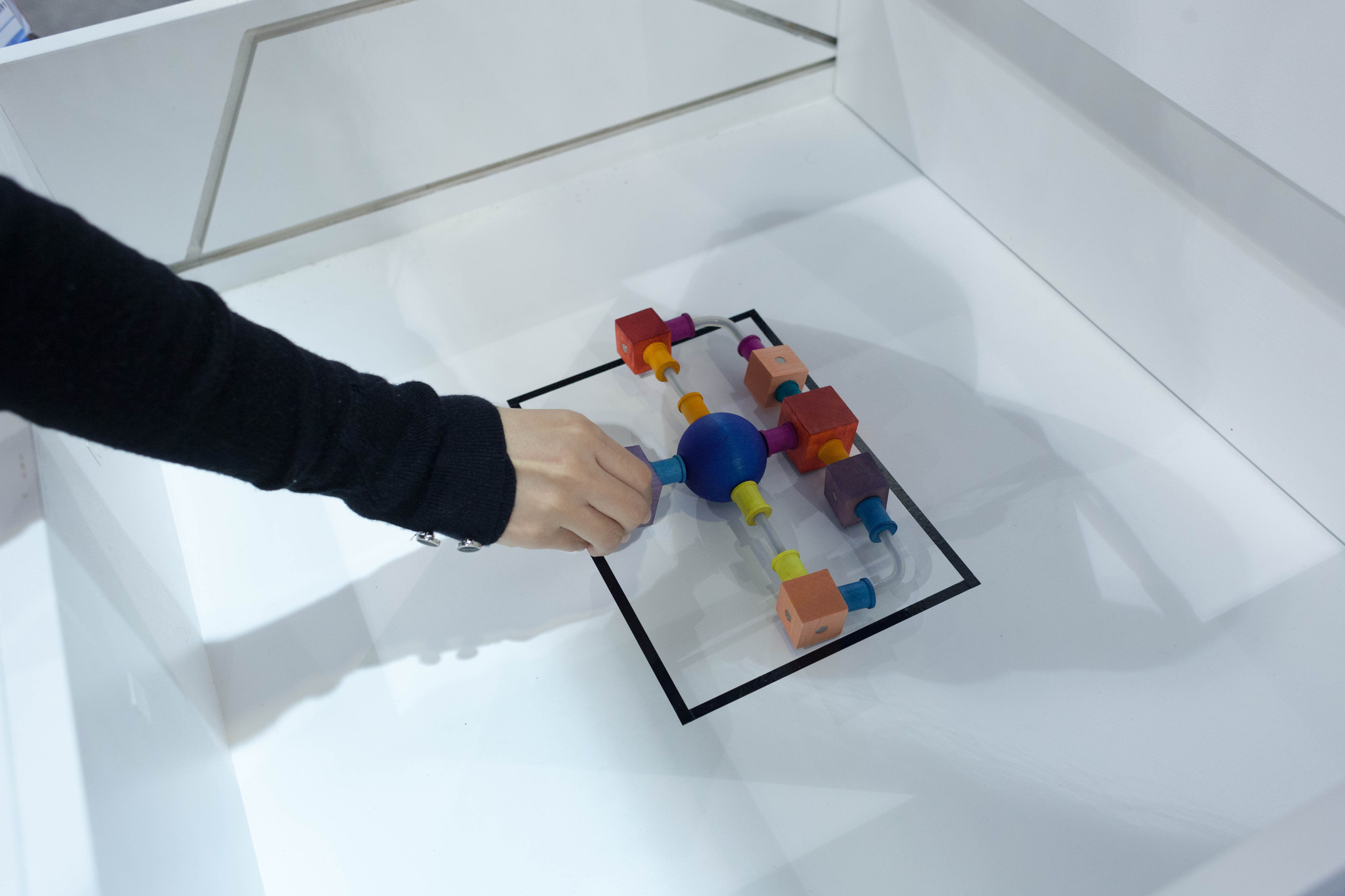 A hand places a 'curio' made of colored wood blocks sits inside a black rectangle.