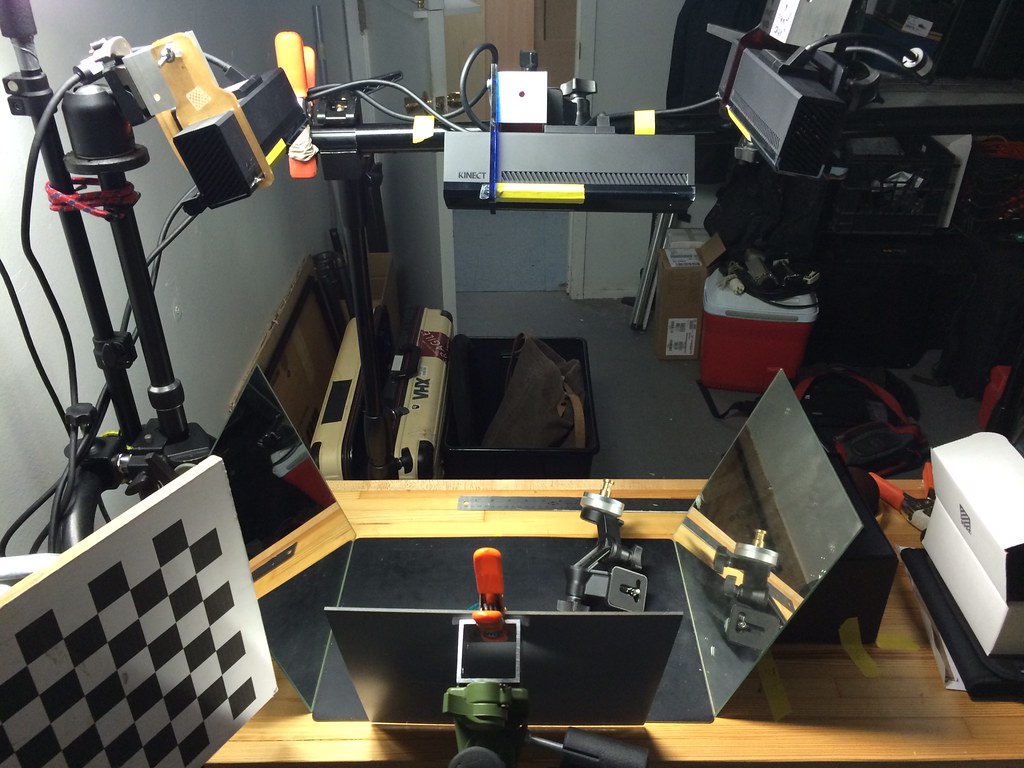 A studio test setup for calibrating three Kinect cameras through angled mirrors, with a checkerboard target and compact experimental capture rig.
