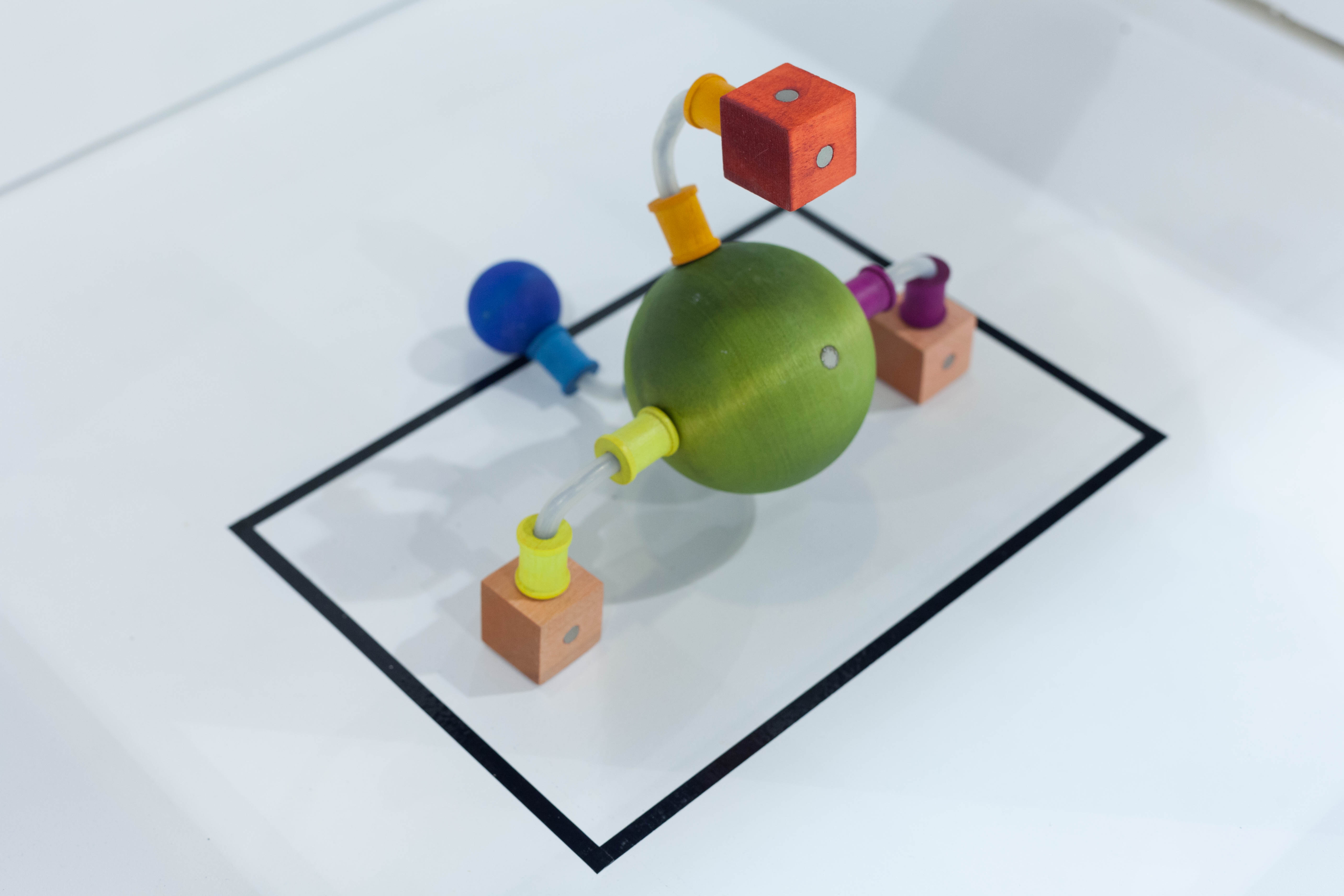 A green wooden sphere connects by clear tubes to red and peach cubes and a smaller blue sphere inside a black display square.