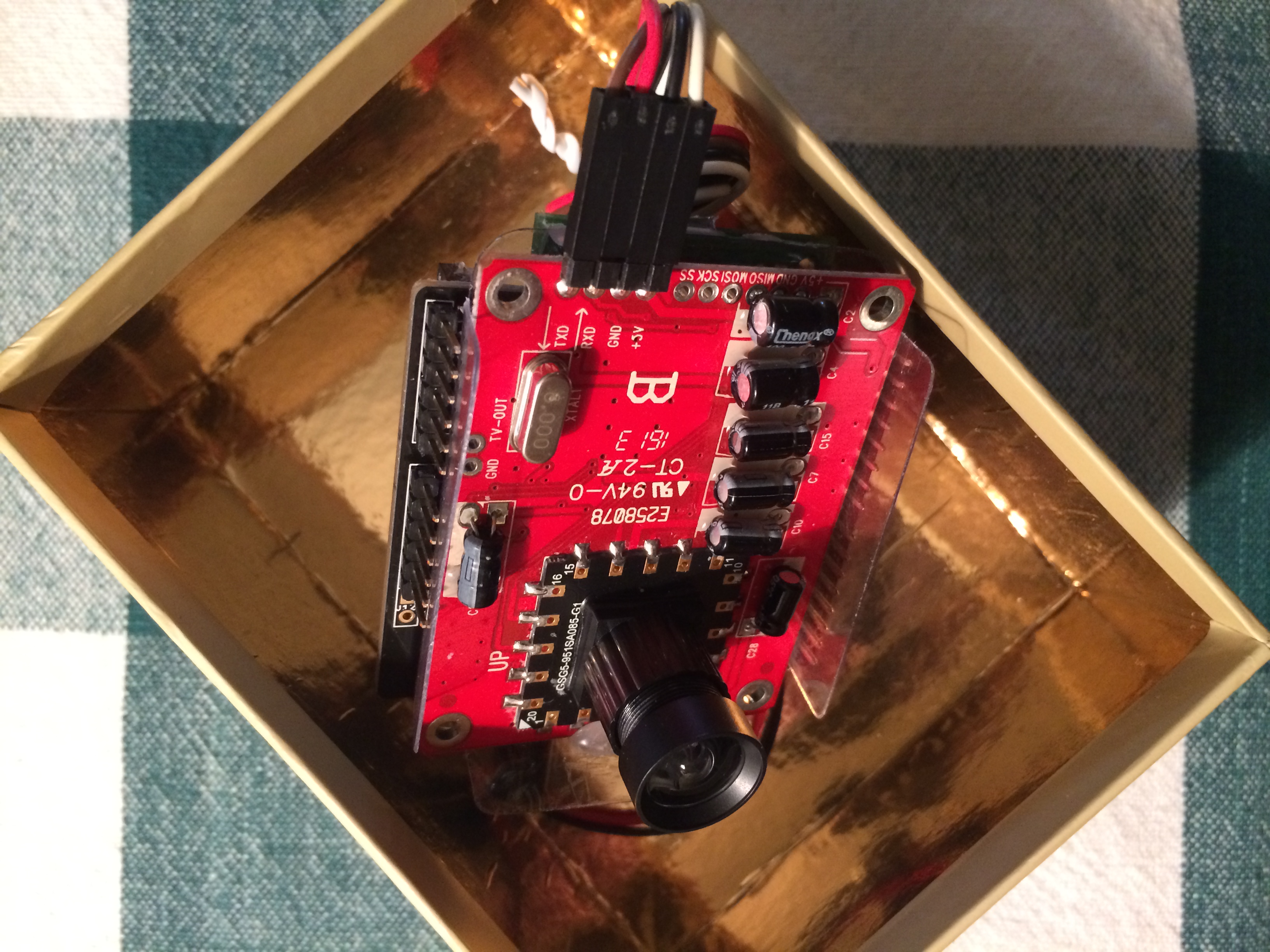 A small red camera circuit board with a lens and exposed components sits inside a copper-lined enclosure.
