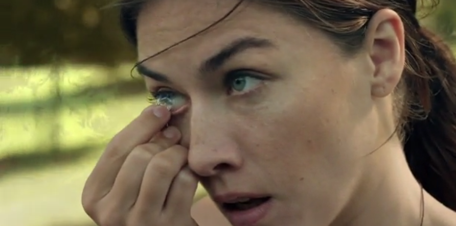 A woman adjusts a transparent contact lens while standing in a sunlit park in Eden 2045.