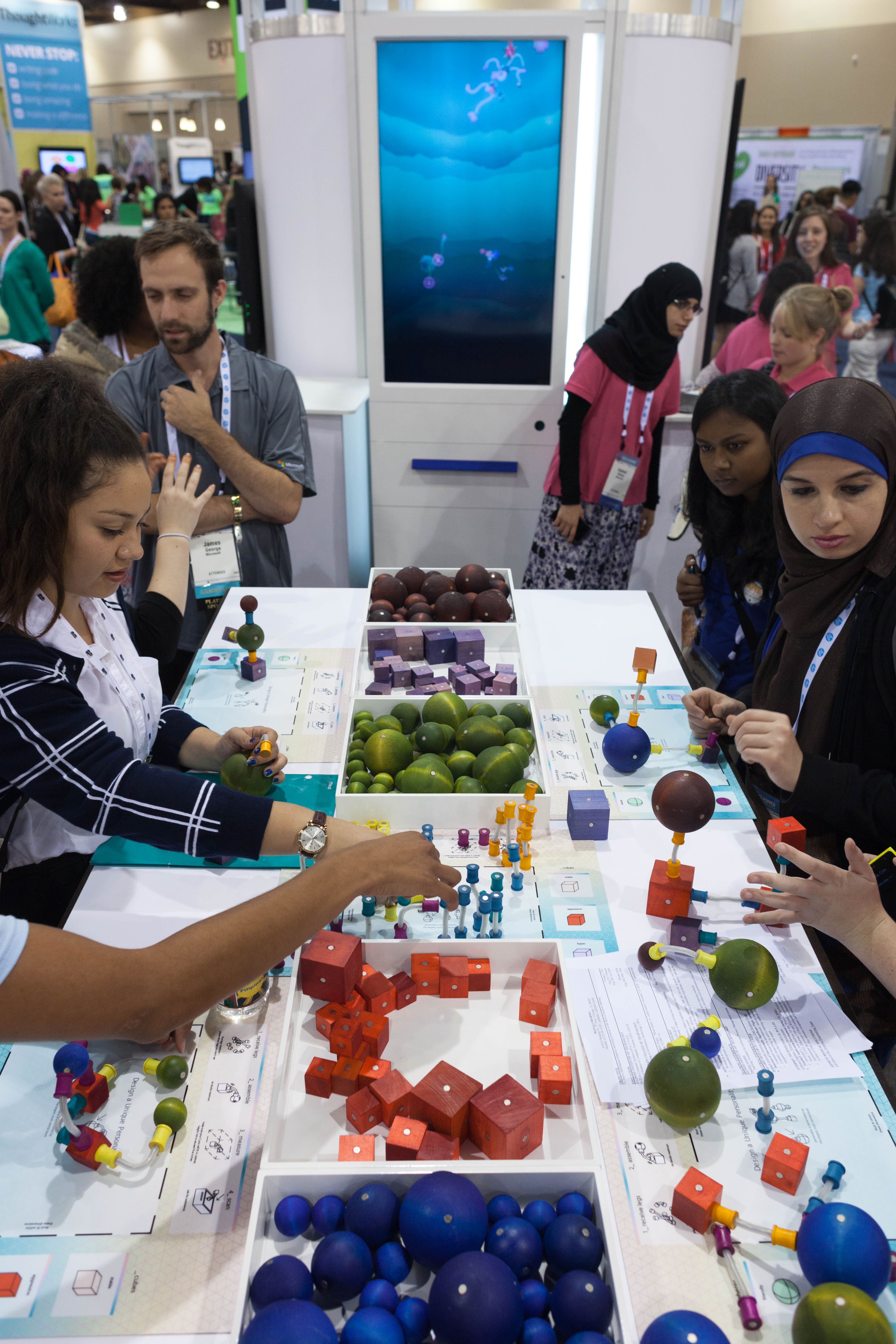 A crowded workshop table holds bins of colorful wooden spheres, cubes, tubes, and pegs as participants build modular Curios around a tall aquarium display.