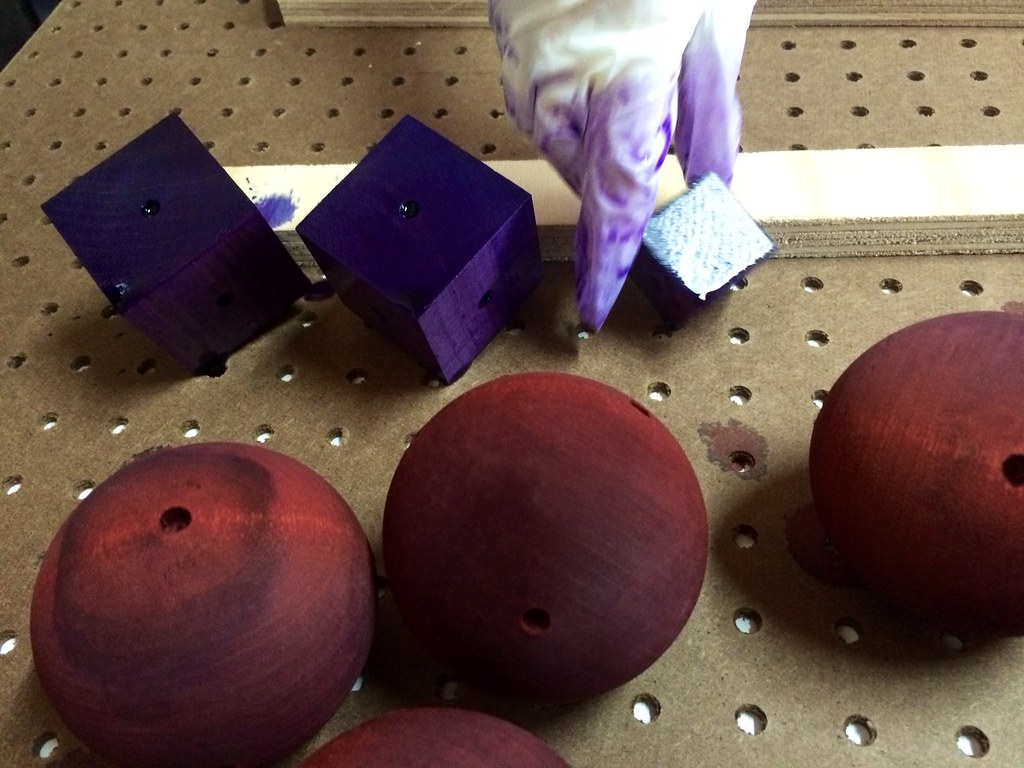 A dye-stained gloved hand works among purple wooden cubes and rounded toy components during fabrication.