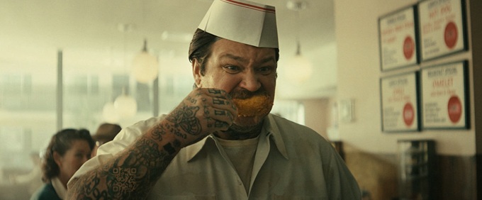 Tattooed man in a paper restaurant hat covering his mouth with one hand.