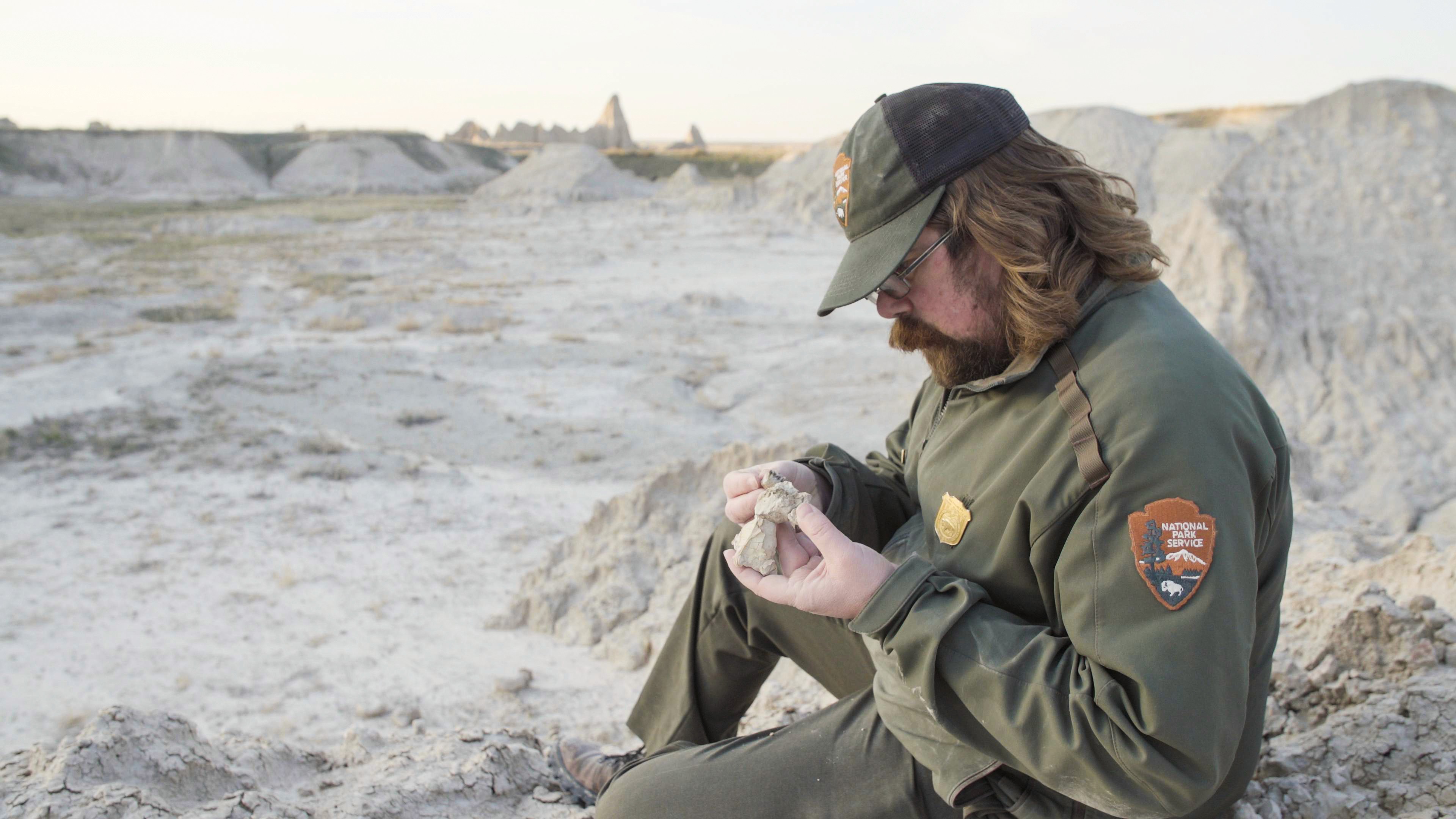 A National Park Service staff member examines material while seated among pale, eroded Badlands formations.