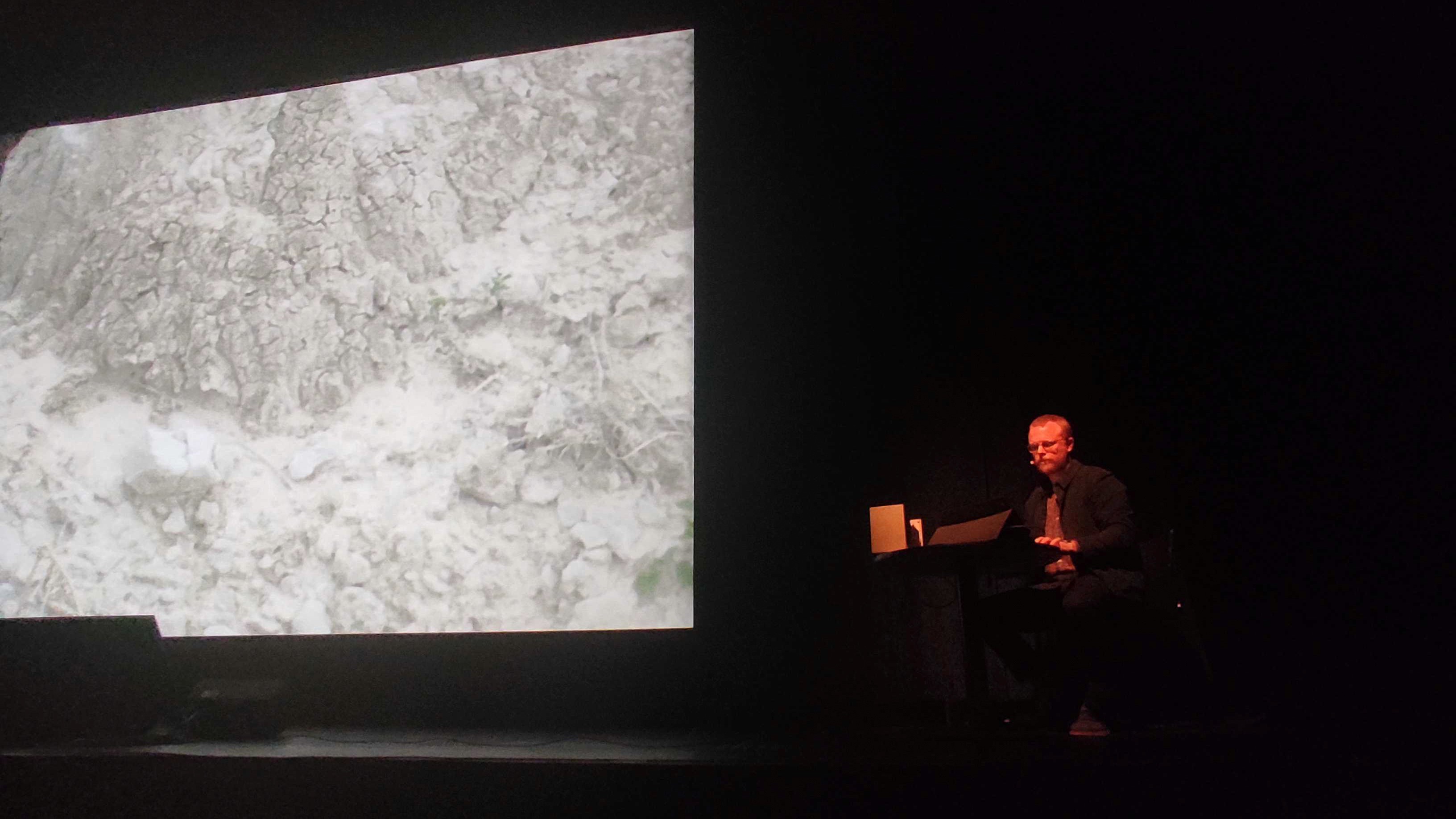 A performer sits beside a large monochrome projection of scanned terrain on a dark stage.