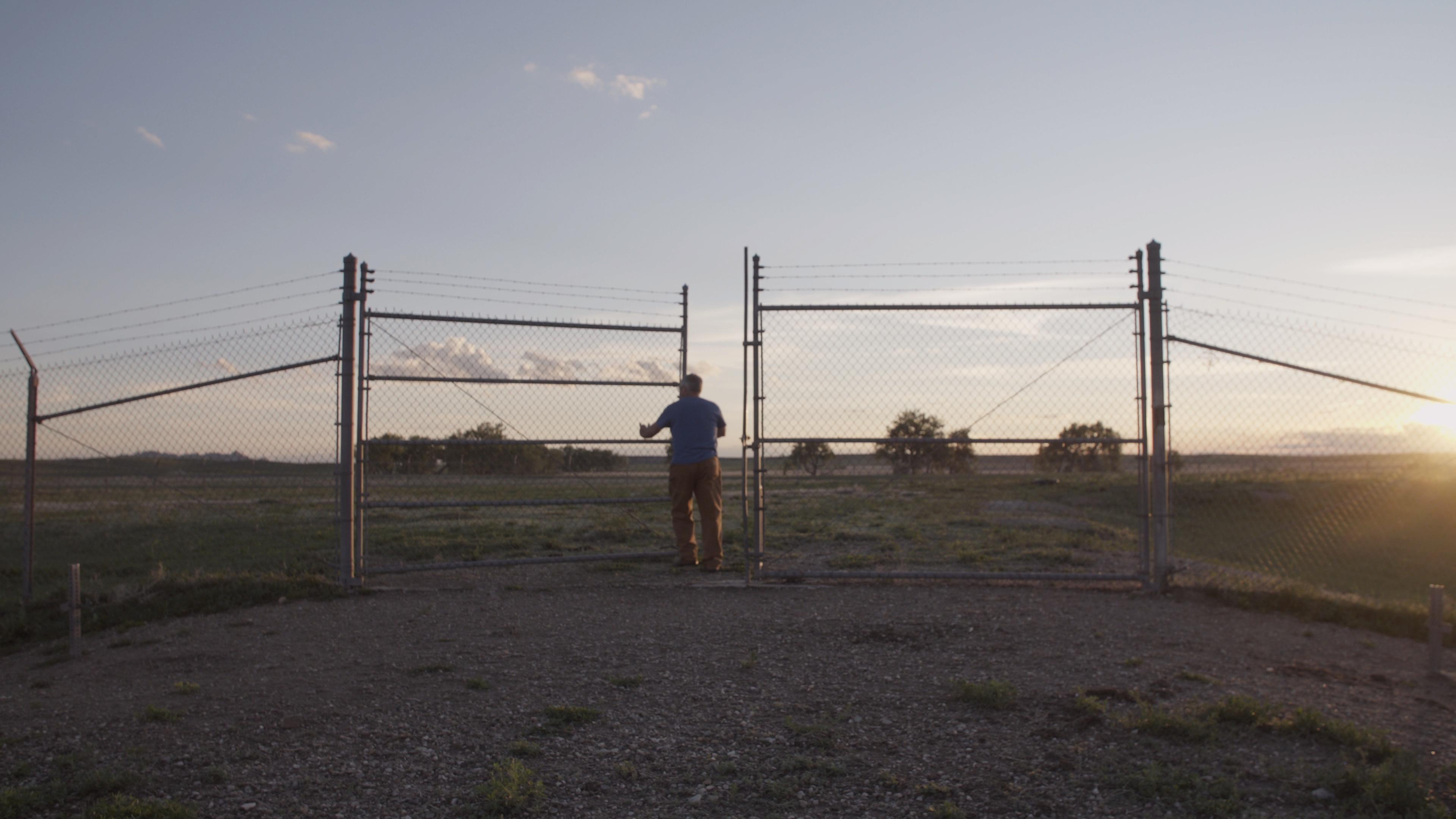 A person opens a wide chain-link gate at the edge of the Badlands as the sun sets behind the fence.