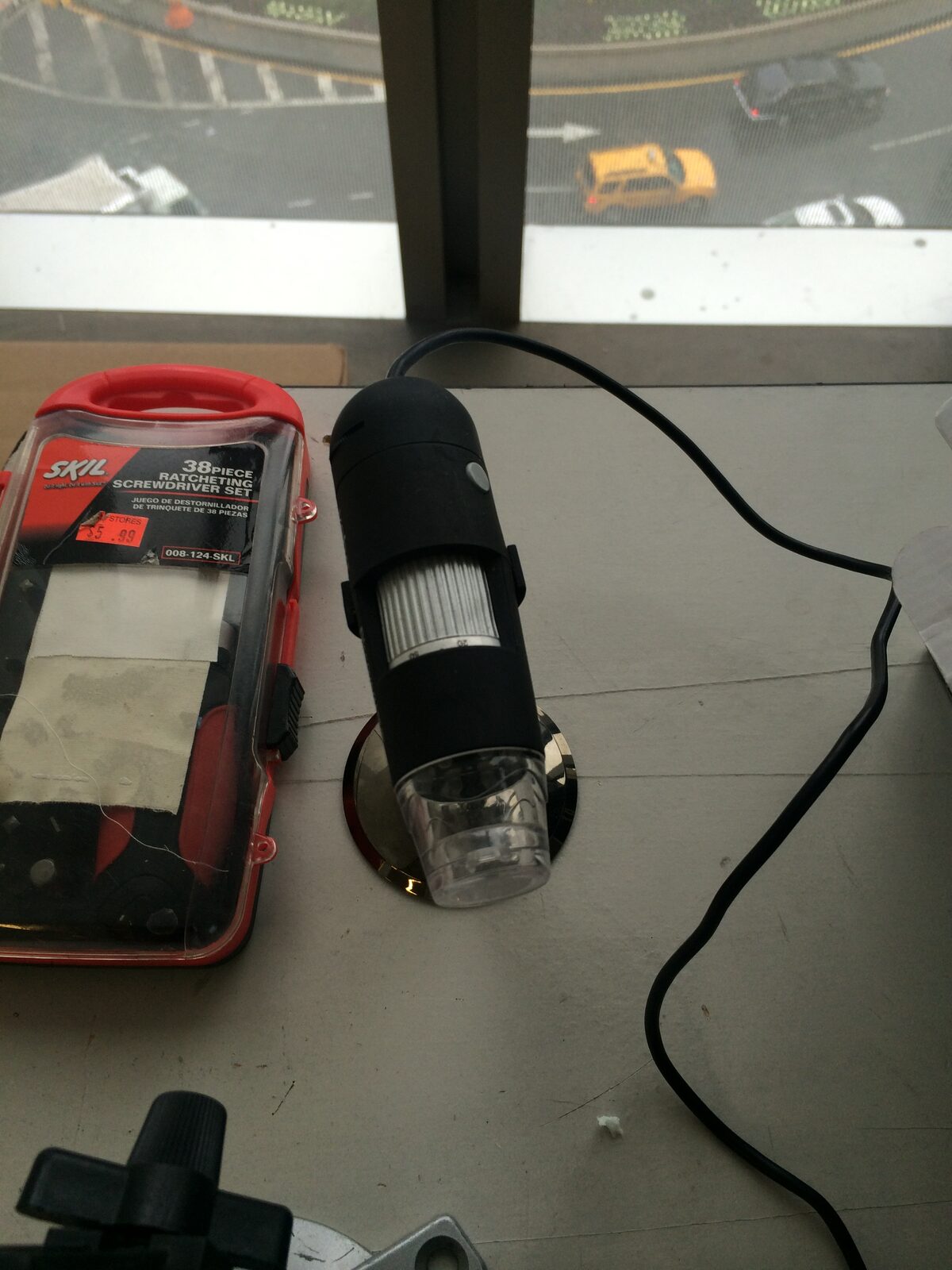 A small USB digital microscope beside a screwdriver set on a workshop table overlooking a city street.