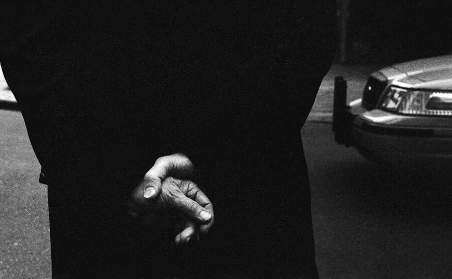 Close black-and-white view of a person's clasped hands beside a parked car.