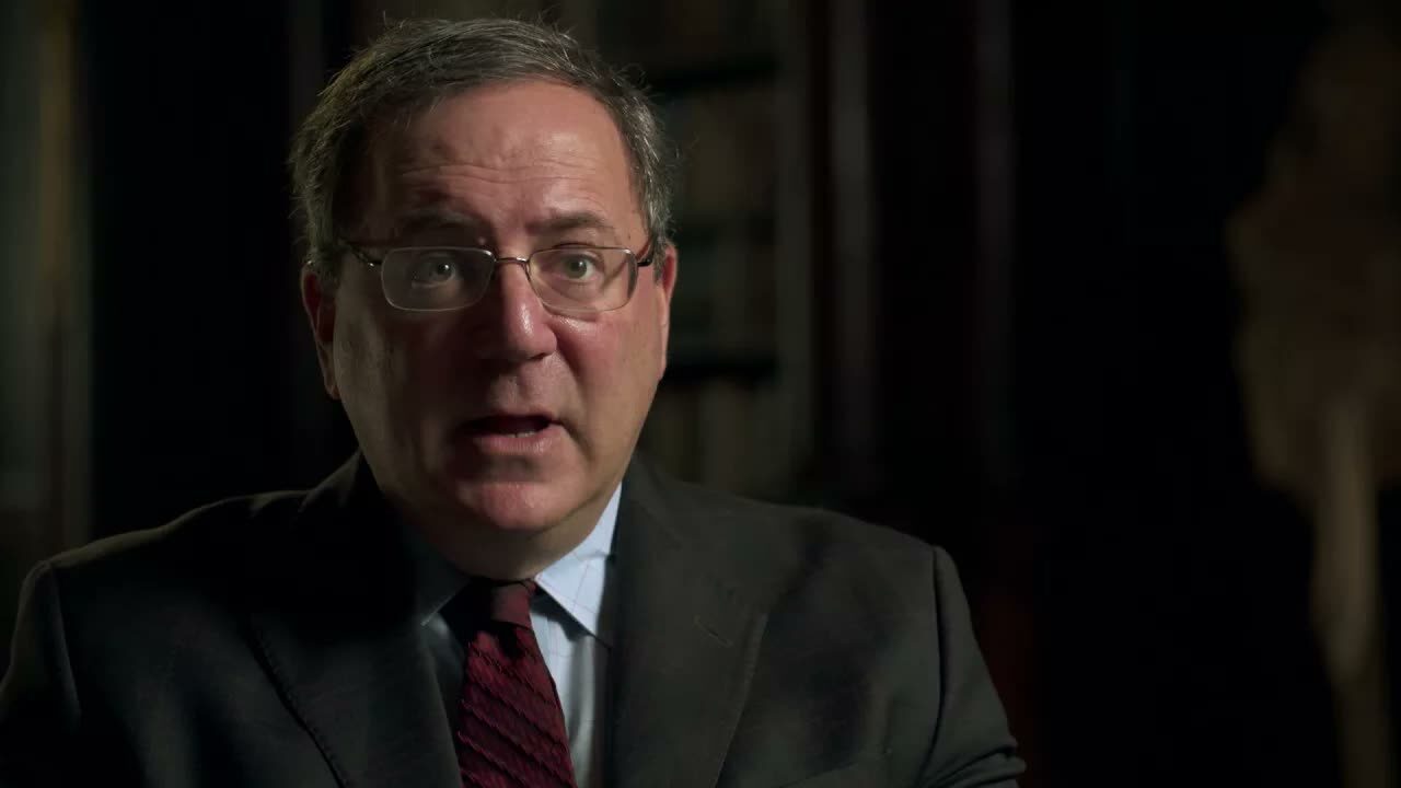 Interview subject in glasses and a suit speaks against a dark background in an official film clip.