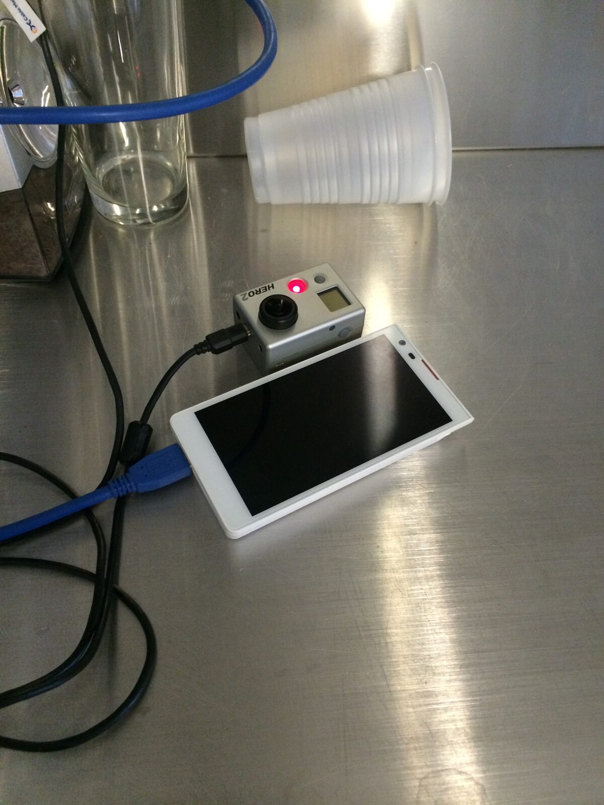 A GoPro Hero2 camera connected by cables beside a white mobile display on a stainless-steel workshop table.