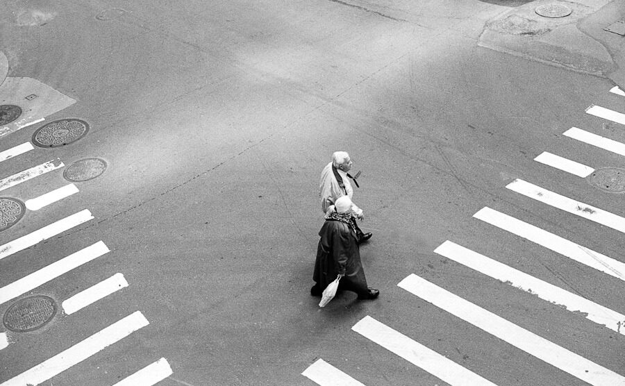 Overhead black-and-white view of a pedestrian crossing a striped intersection.