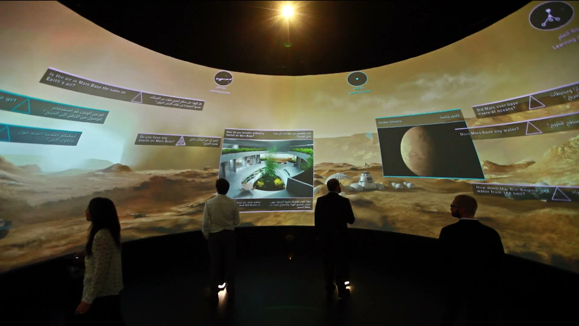 Visitors stand inside a dark circular room surrounded by a projected Martian landscape, bilingual questions, diagrams, and responsive lesson panels.